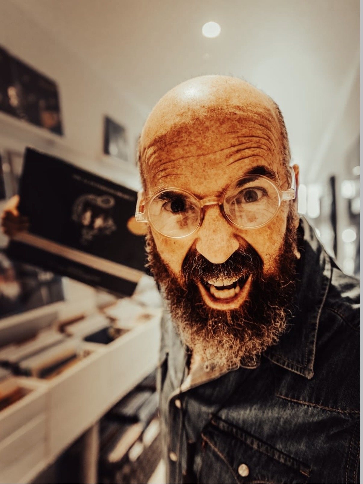 A grinning record dealer in round glasses holds a sleeve up to the camera inside Slow Life Records
